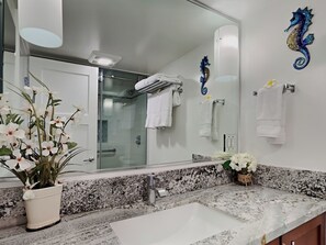 Un baño con lavamanos blanco, encimera de granito y un espejo grande. Hay toallas colgadas en la pared y una planta en maceta con flores blancas sobre la encimera.