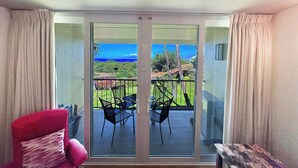 A room with a sliding glass door leading to a balcony with a view of the ocean and a garden. There is a red chair and a small table on the balcony. - Luxurious Kihei Remodeled 2BR Condo Sweeping Ocean Views Near Keawakapu (Kihei)
