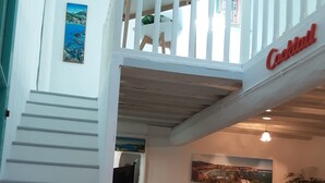 Un escalier menant à un balcon orné d’une enseigne indiquant « Cocktails », un canapé avec des coussins aux couleurs vives et un tableau accroché au mur.