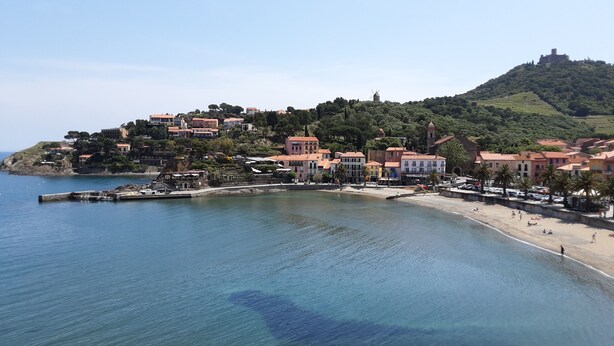 Une ville côtière avec des bâtiments construits sur une colline, une plage et une mer d’un bleu limpide.