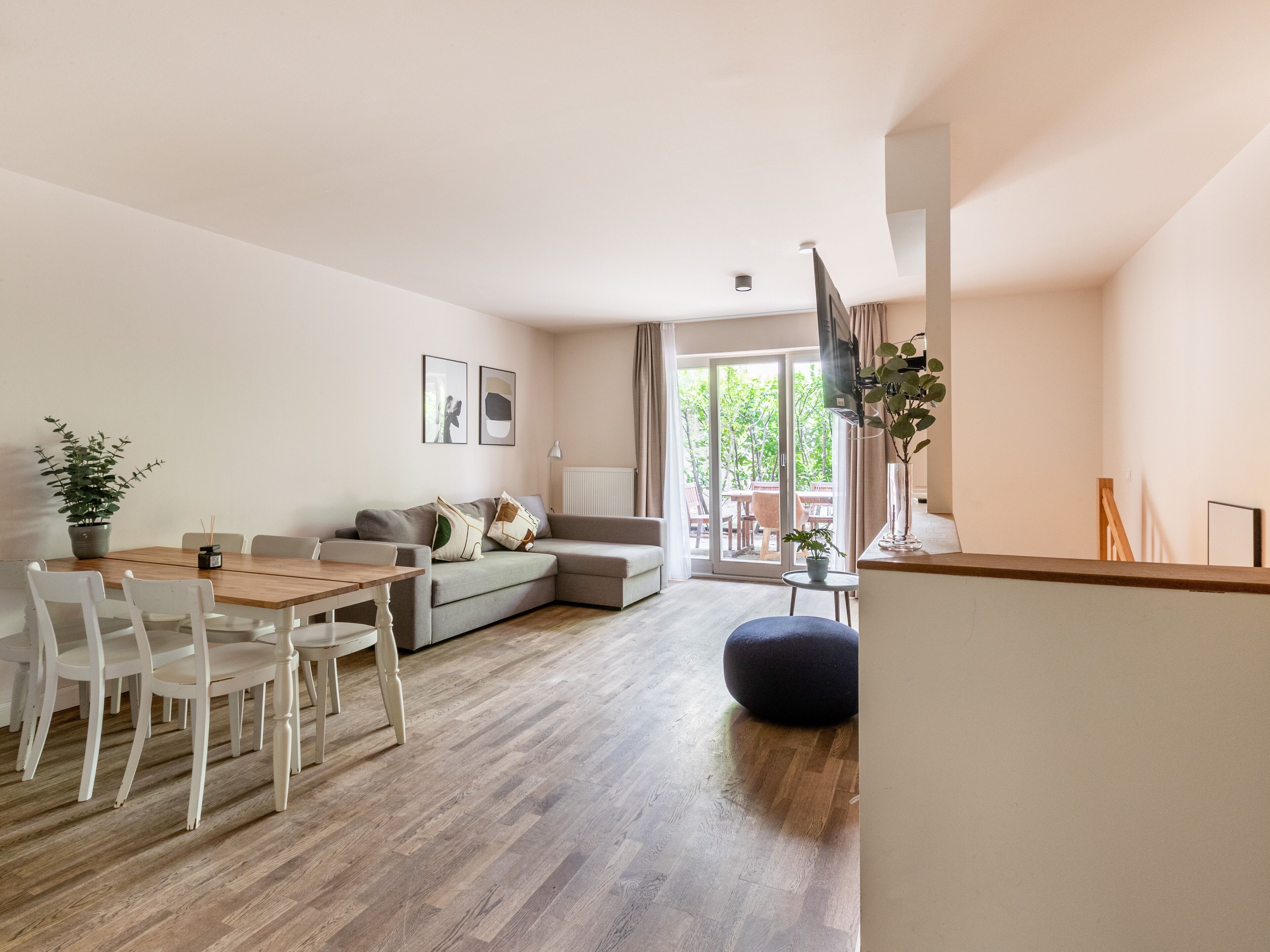 A modern living room with a wooden dining table, white chairs, a grey sofa, and a blue ottoman. There is a TV mounted on the wall, a potted plant, and a window with a view of greenery outside.