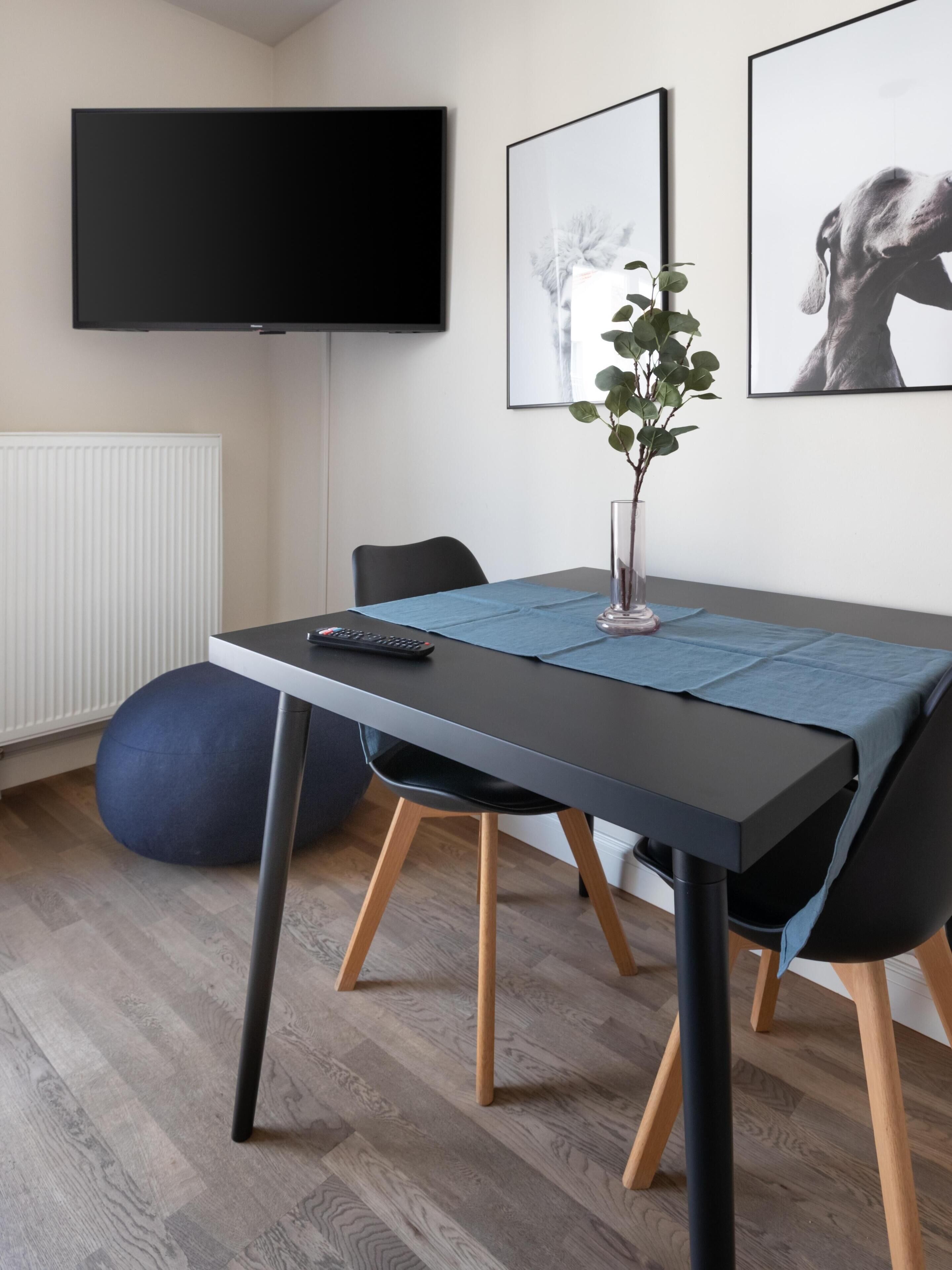 A modern dining area with a rectangular table, black chairs, a blue table runner, a vase with a plant, and a flat-screen TV mounted on the wall.