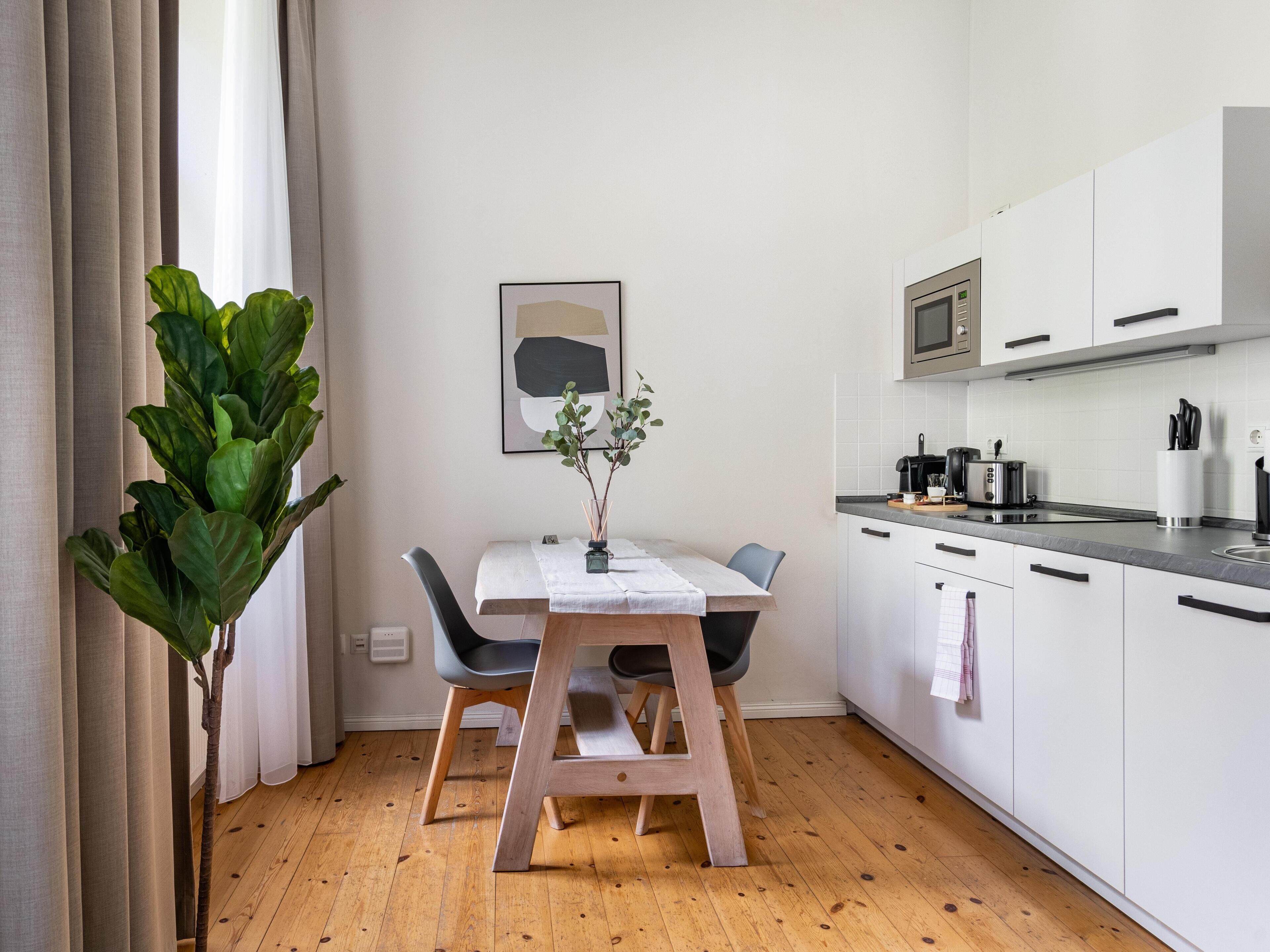 Moderni keittiö, jossa on valkoiset kaapit, puinen ruokapöytä ja ruukussa oleva kasvi.