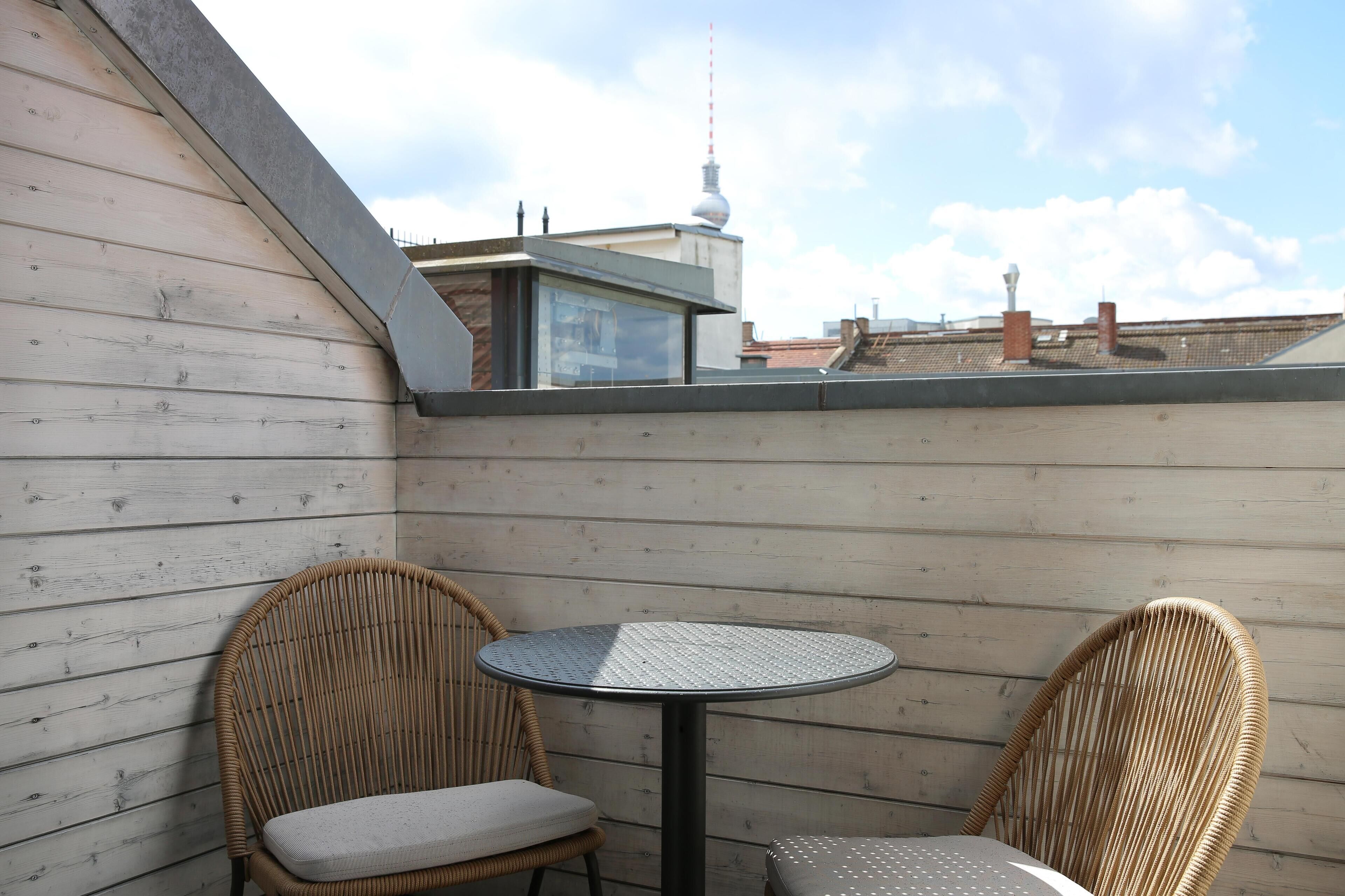 Een terras op het dak met twee rieten stoelen en een kleine ronde tafel, wanden van houten panelen en een uitzicht over een stadsgezicht inclusief een toren.