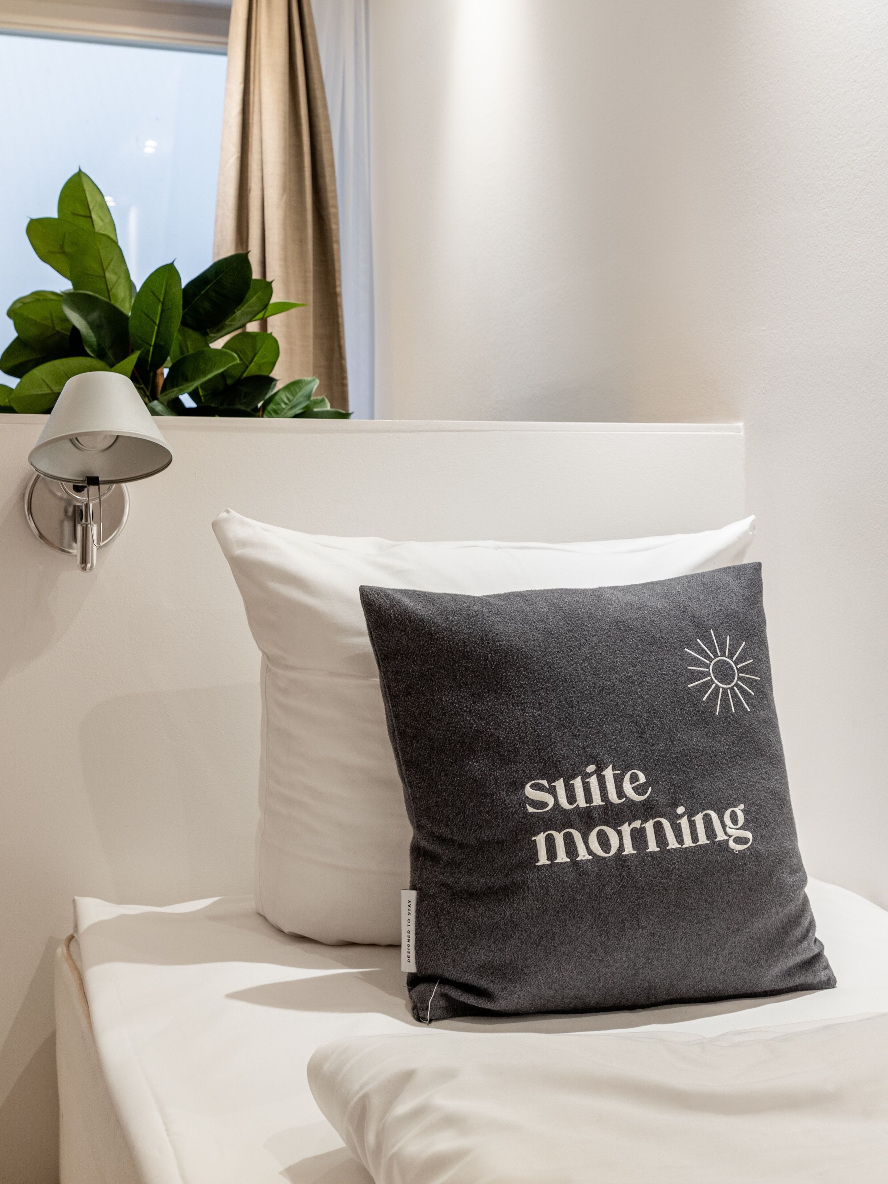 Un lit bien fait, avec un oreiller orné de l’inscription « bonne matinée » et une lampe fixée au mur.