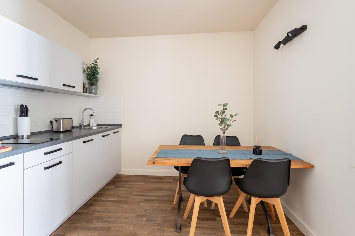Une cuisine compacte avec des meubles blancs, un plan de travail noir et une table à manger en bois, assortie de chaises noires.