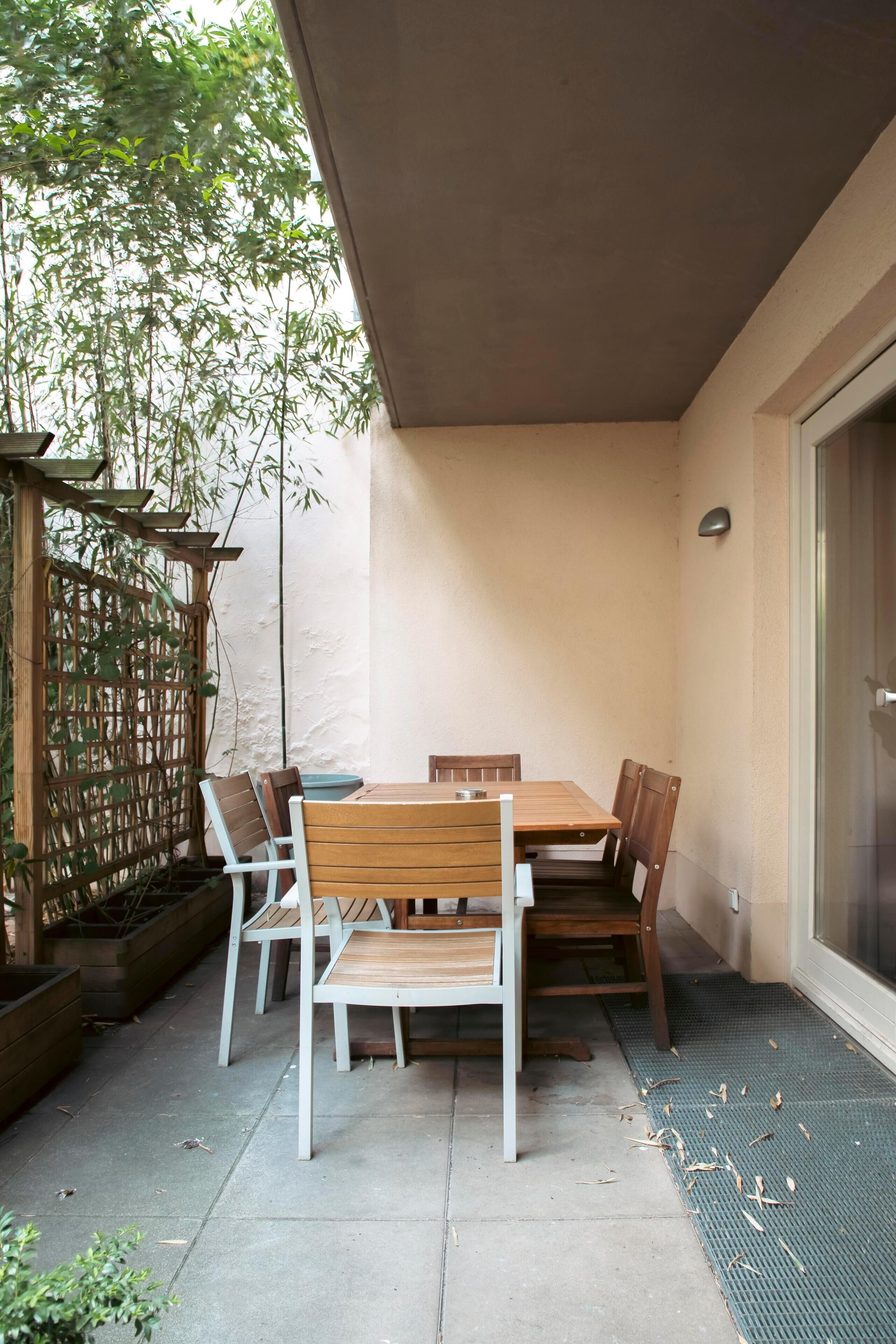 A covered patio with a wooden table and chairs, a bamboo plant, and a glass door.