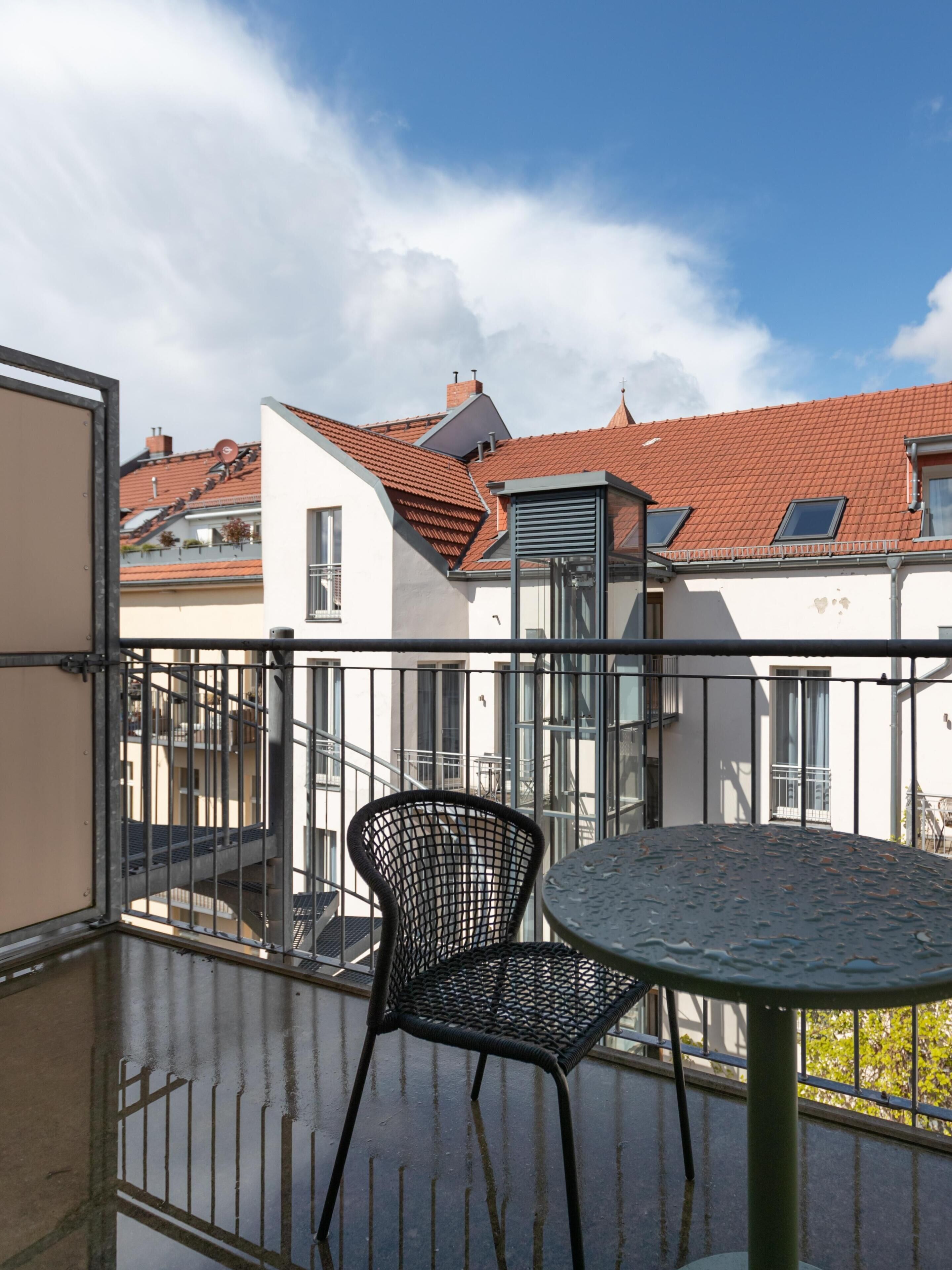 En balkon med et rundt bord og en sort stol, med udsigt over et boligområde med røde tegltage og hvide vægge.