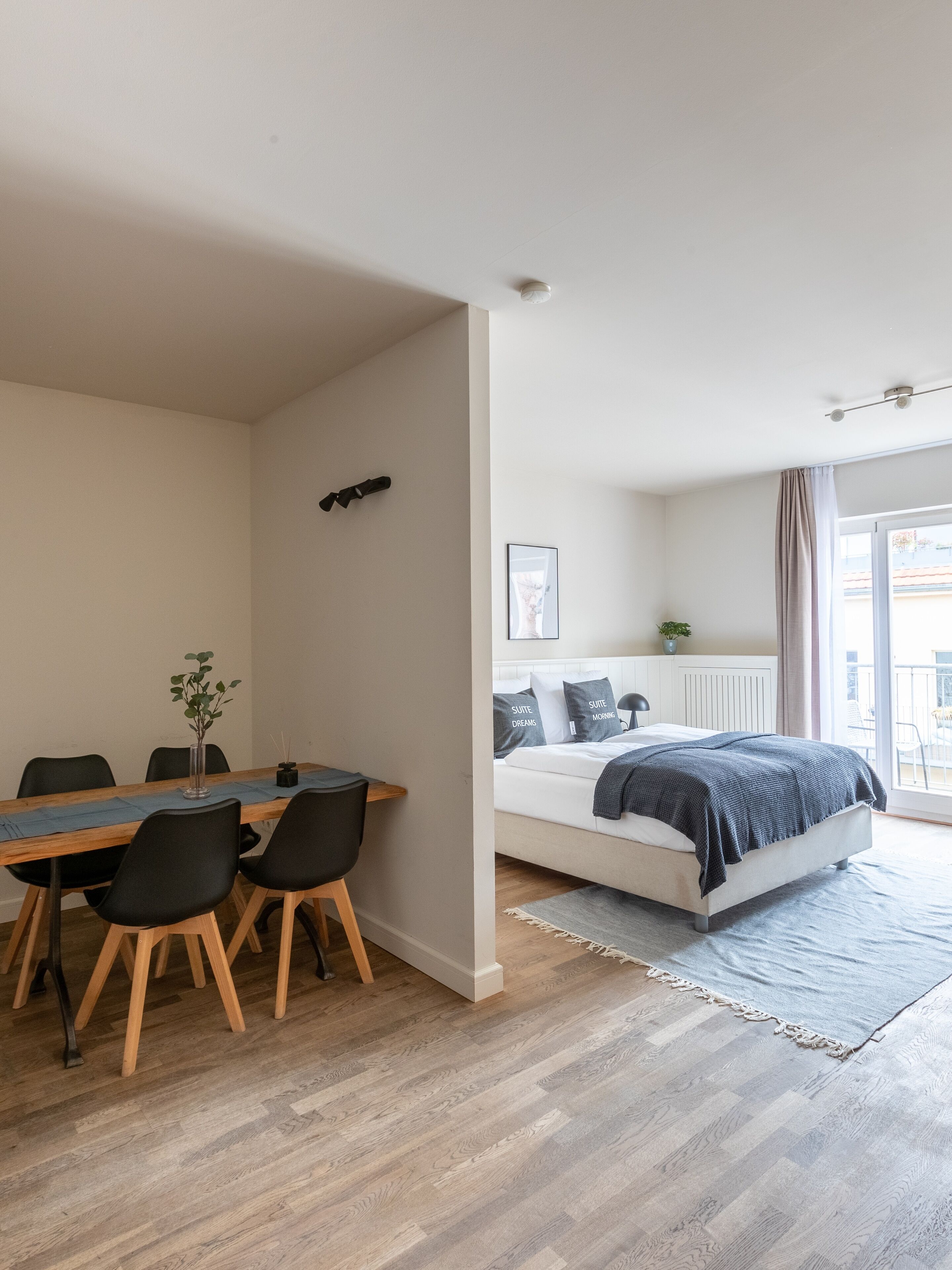 A modern room with a wooden dining table, black chairs, a bed with a grey blanket, and a large window.