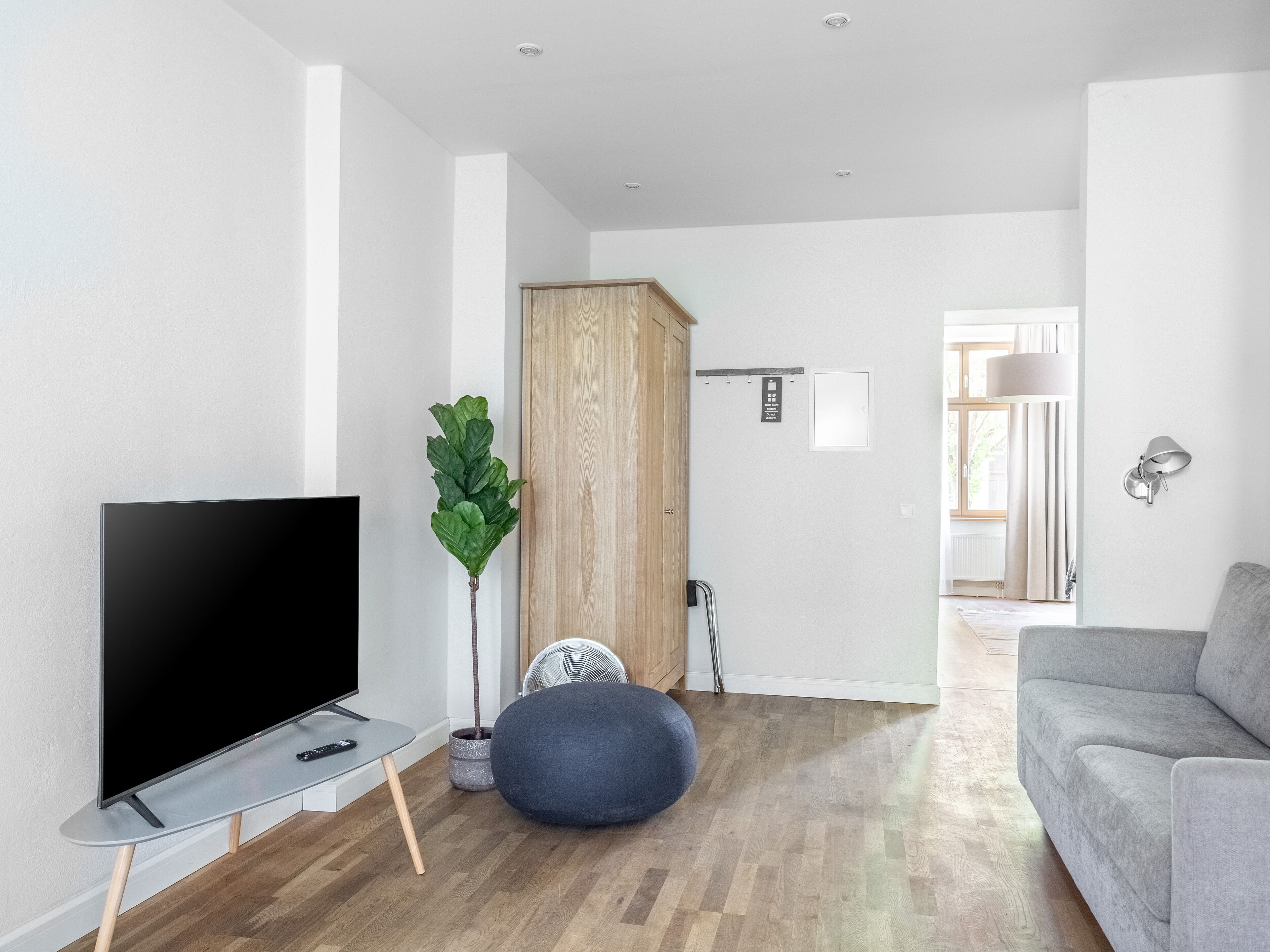 Uma sala de estar moderna com uma TV de tela plana, um sofá cinza, um puff azul, um armário de madeira e uma planta em vaso.