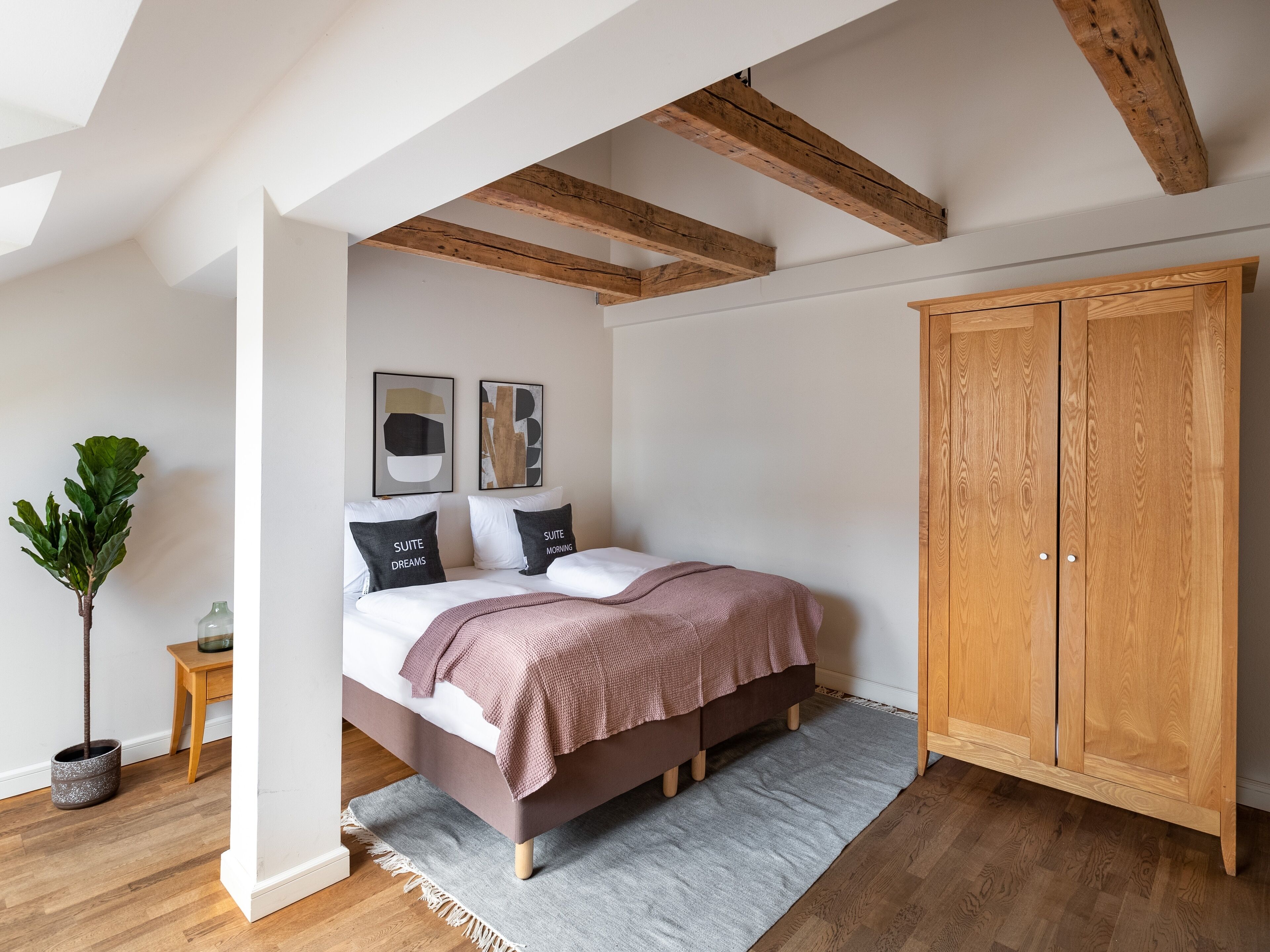 A bedroom with a bed, wooden beams, a wardrobe, and two framed artworks on the wall.