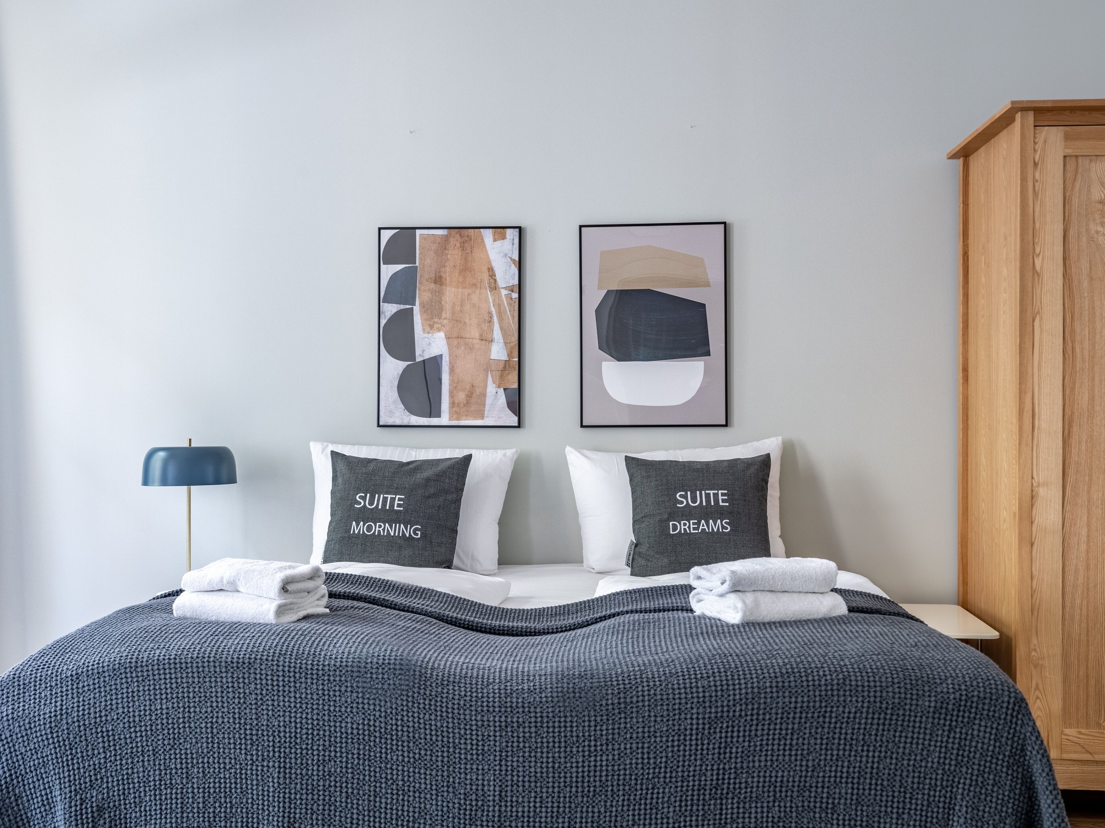 A neatly made bed with two pillows labeled "SUITE MORNING" and "SUITE DREAMS," a blue floor lamp, and abstract art on the wall.