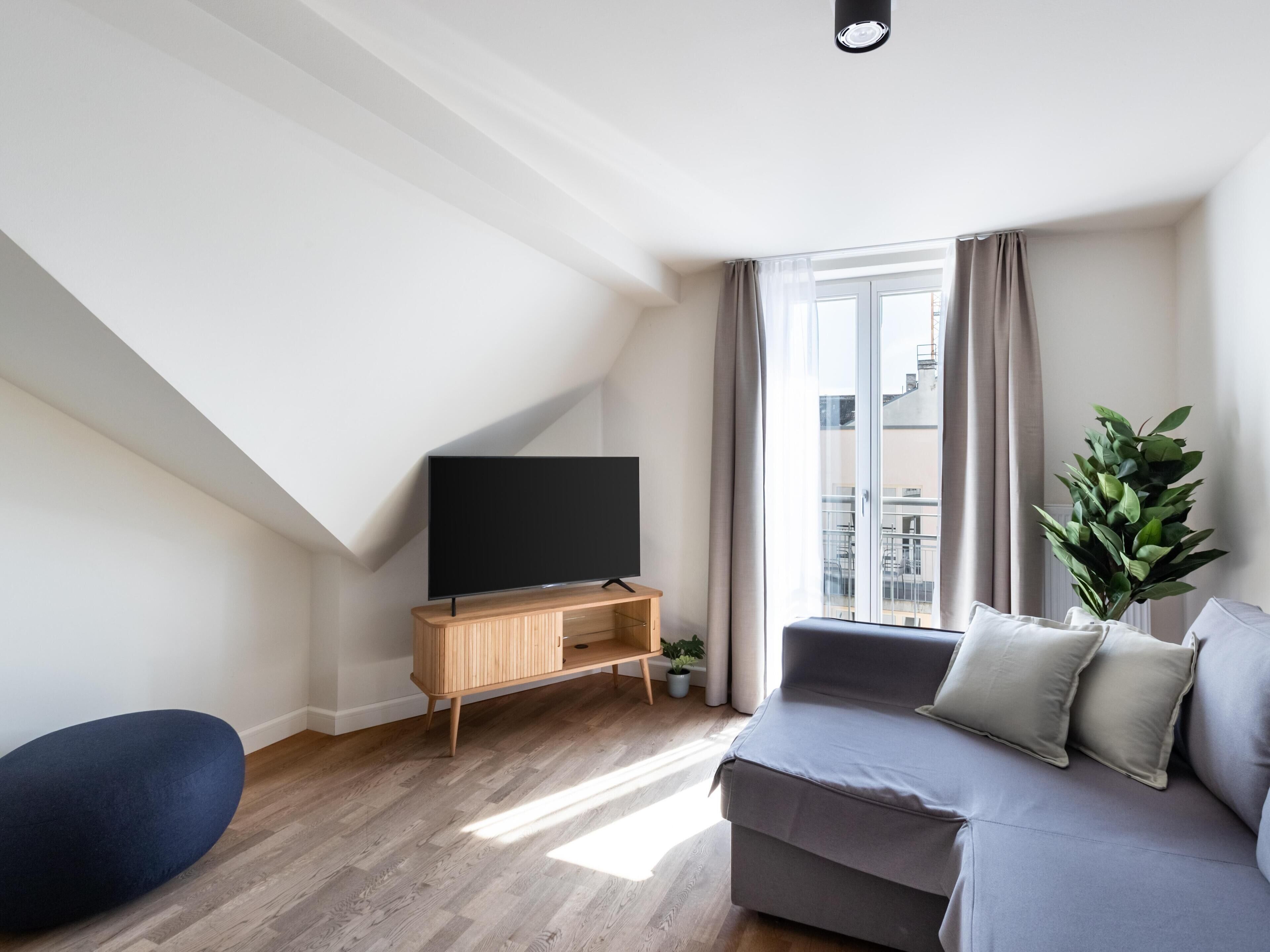 A modern living room with a flat-screen TV on a wooden stand, a grey sofa with cushions, a blue ottoman, and a potted plant.