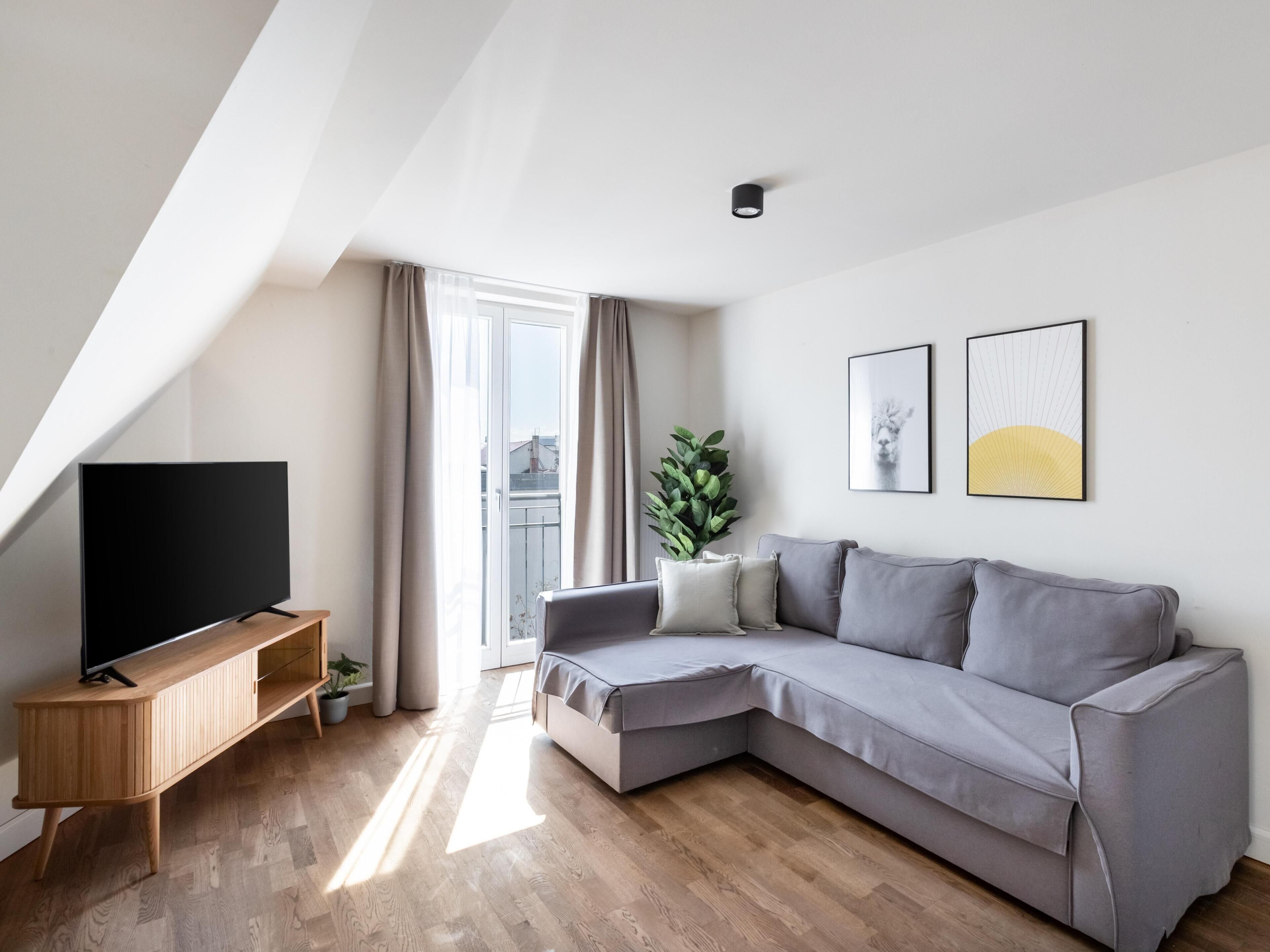 Ein modernes Wohnzimmer mit einer grauen Eckcouch, einem Flachbildfernseher auf einer Holzkonsole, einem Blumentopf und zwei gerahmten Kunstwerken an der Wand.