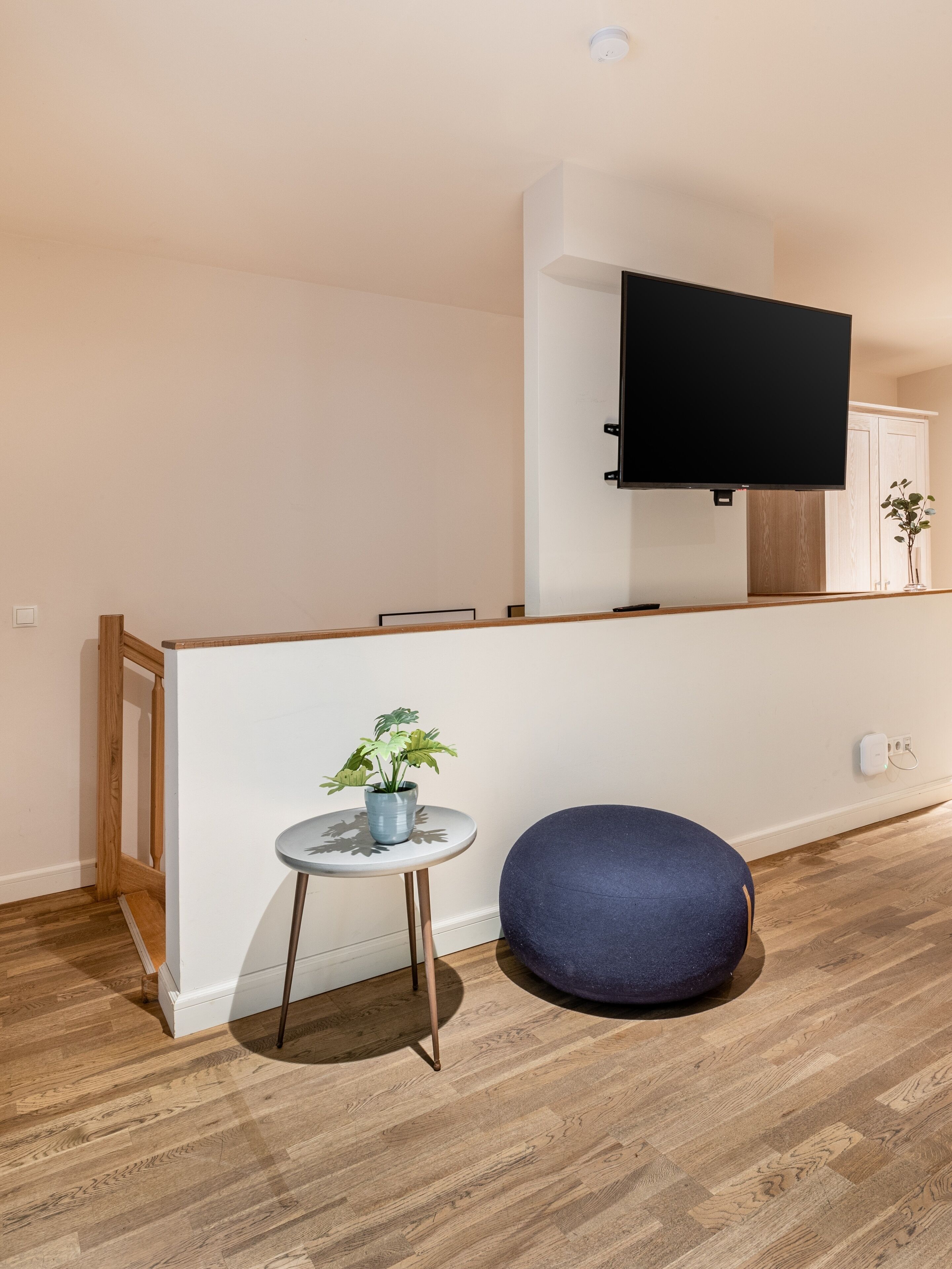 Ein modernes Wohnzimmer mit Holzboden, einem blauen runden Hocker, einem kleinen Tisch mit Pflanze und einem an der Wand montierten Fernseher.