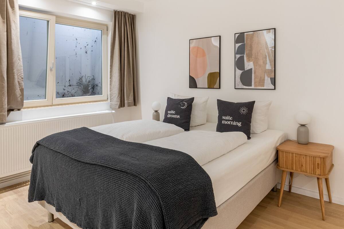 Une chambre à coucher comprenant un lit, une table de chevet, une fenêtre avec des rideaux et deux tableaux abstraits accrochés au mur.
