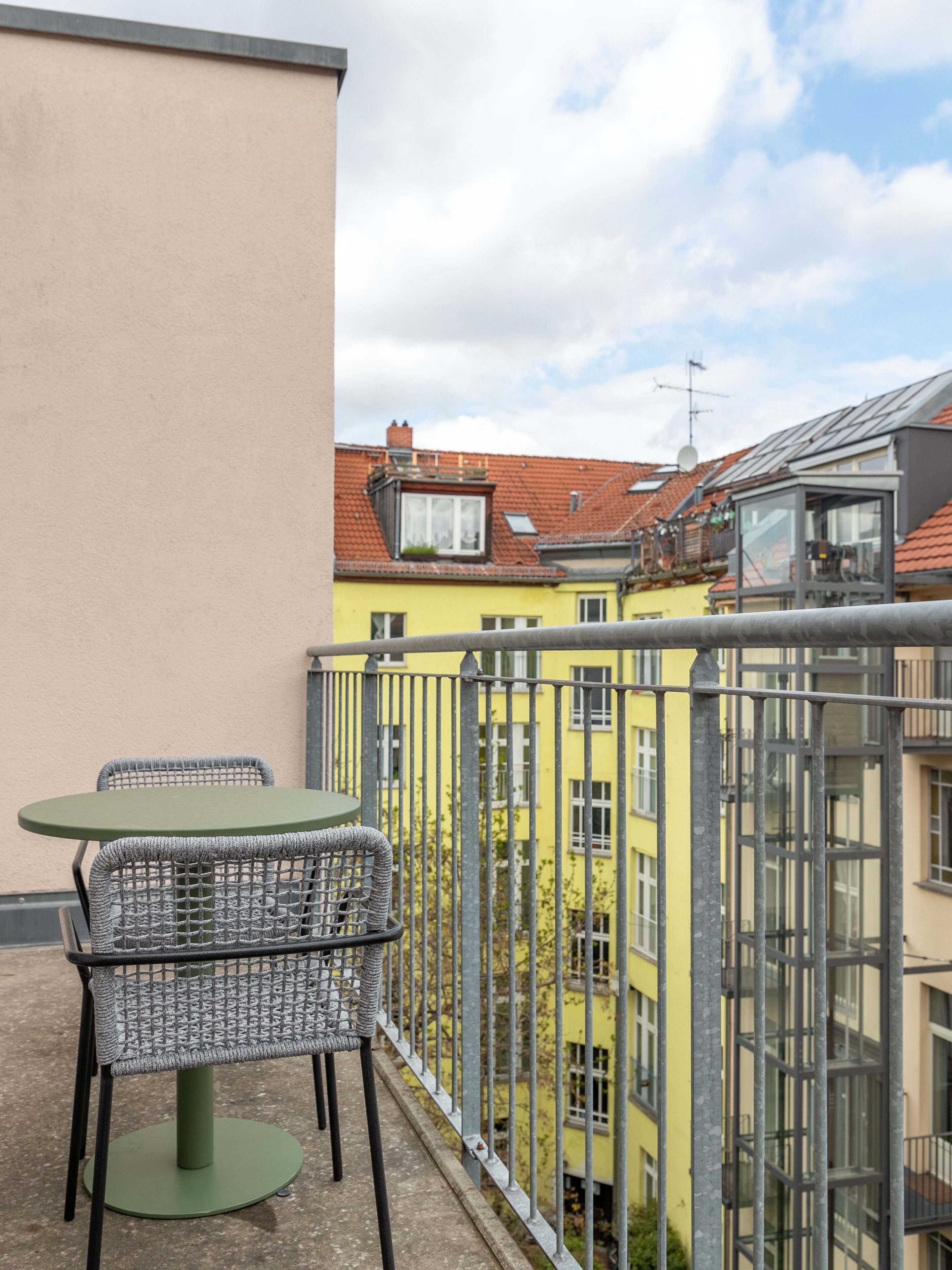 A balcony with a table and chairs, overlooking a row of yellow buildings with red-tiled roofs.