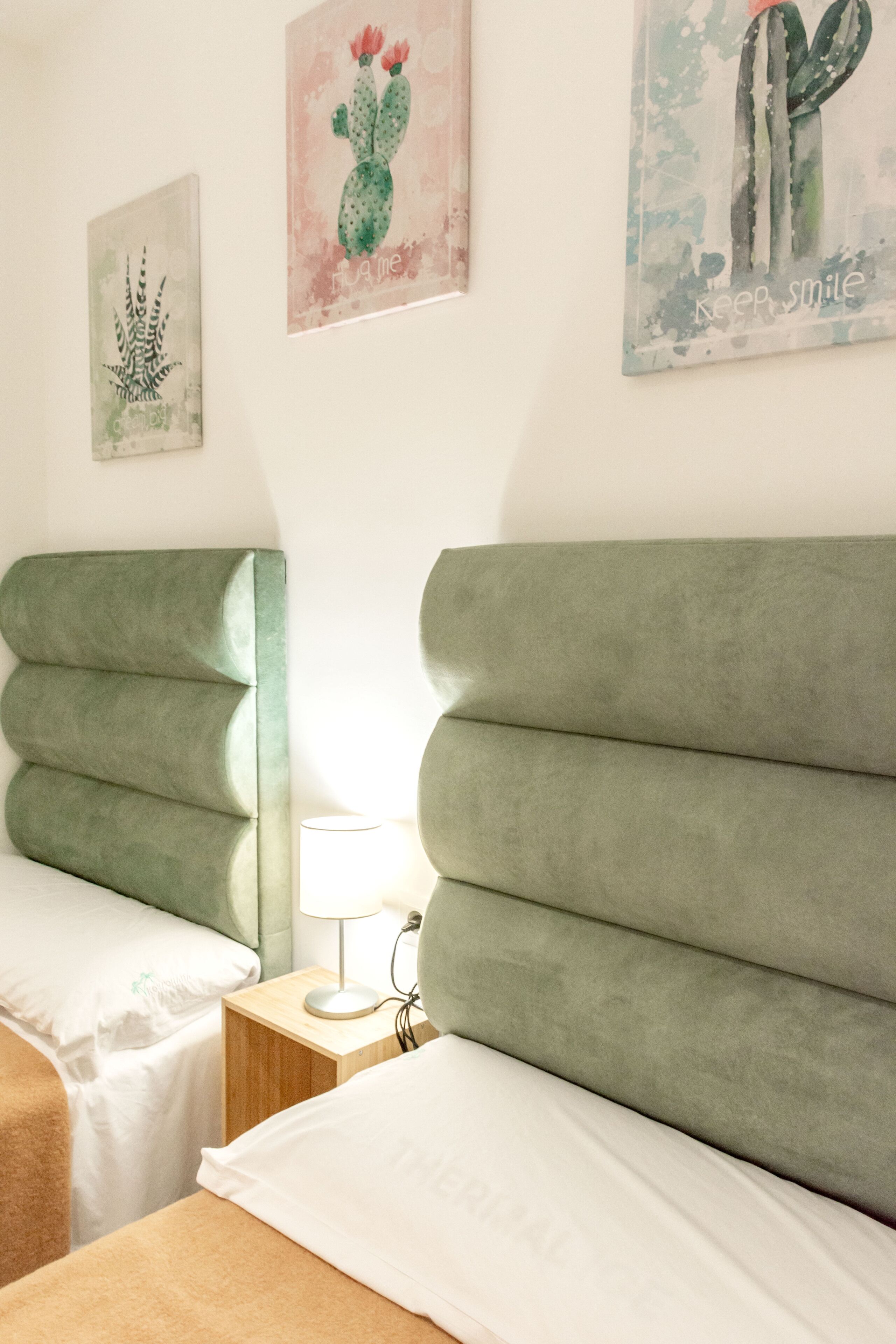 A room with two beds, a nightstand with a lamp, and wall art featuring cacti.