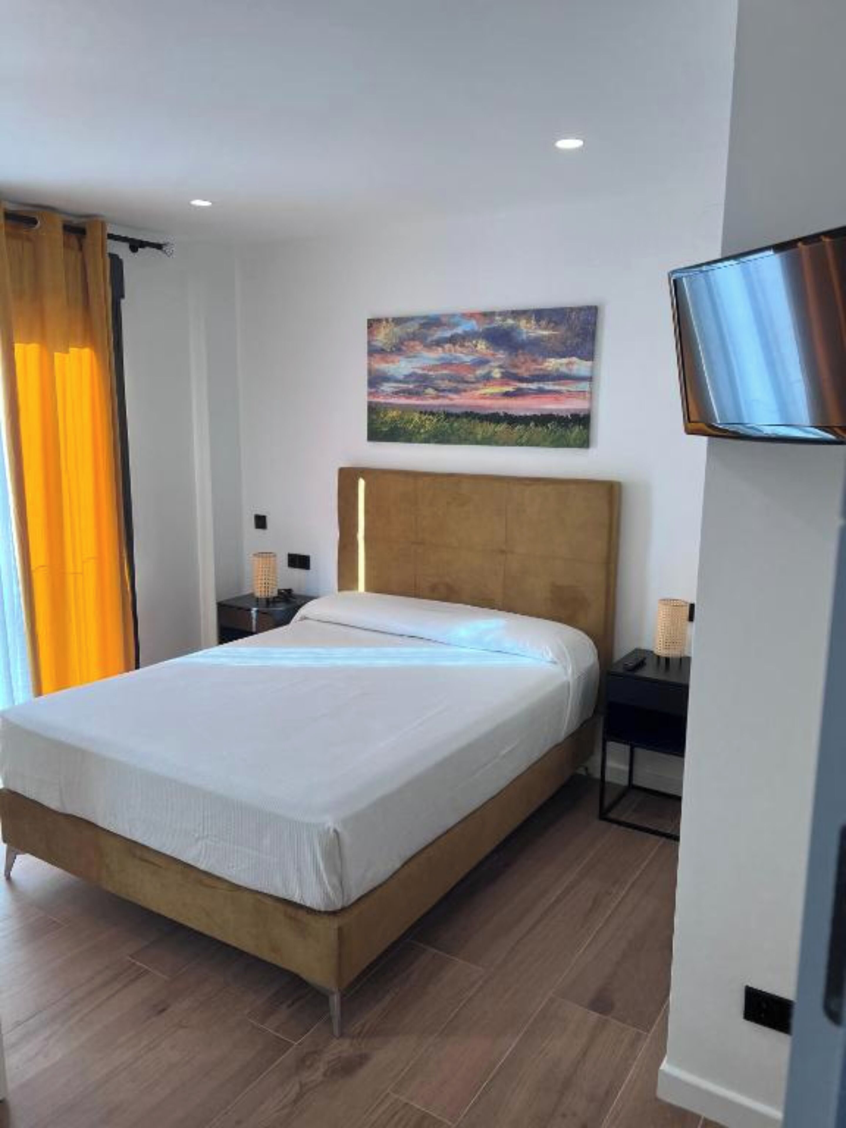 Ein modernes Hotelzimmer mit einem hölzernen Bett, einem Gemälde darüber, einem an der Wand montierten Fernseher und einem Nachttisch mit Lampe.