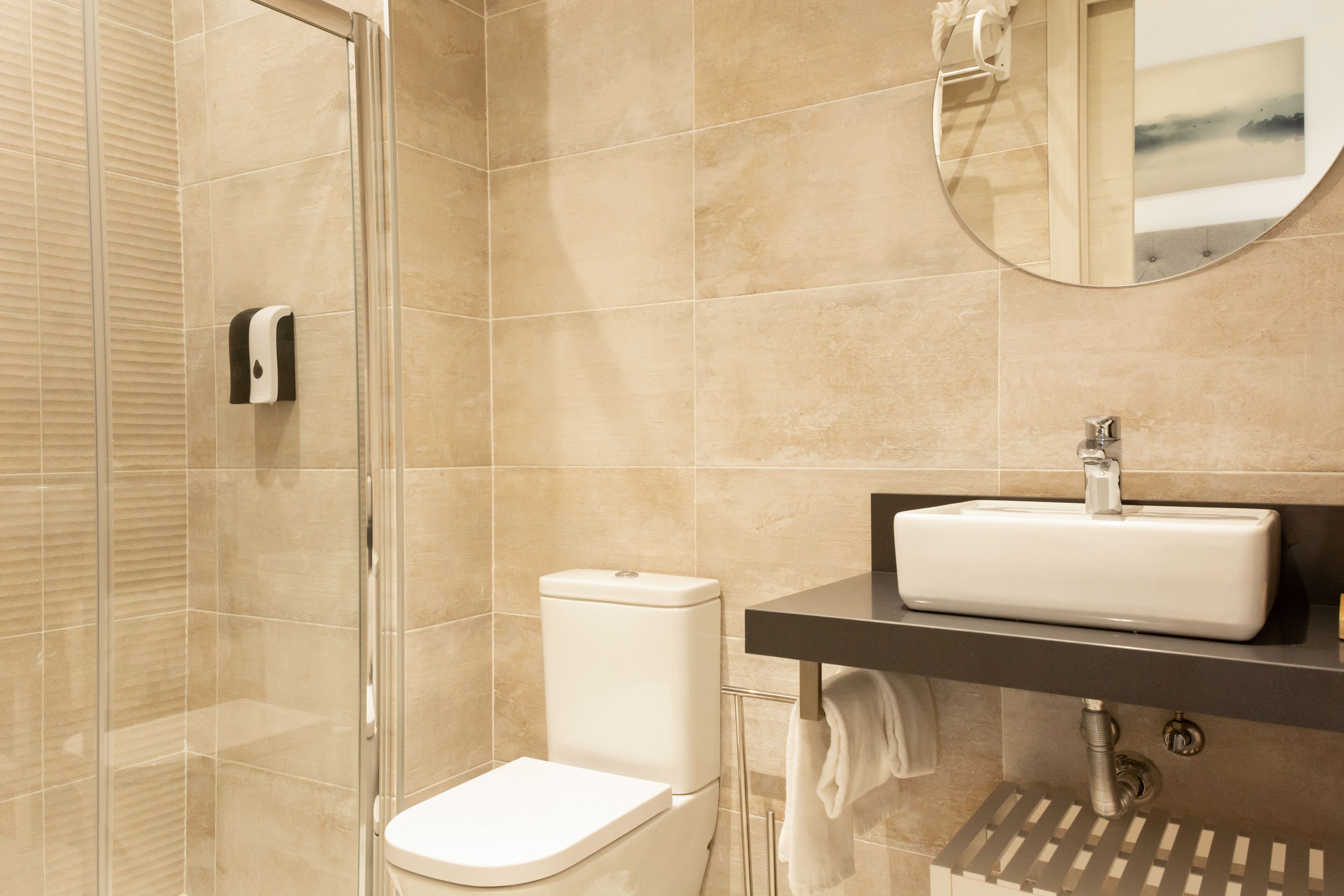 A modern bathroom with a glass shower enclosure, a white toilet, a sink with a mirror above it, and beige tiled walls.