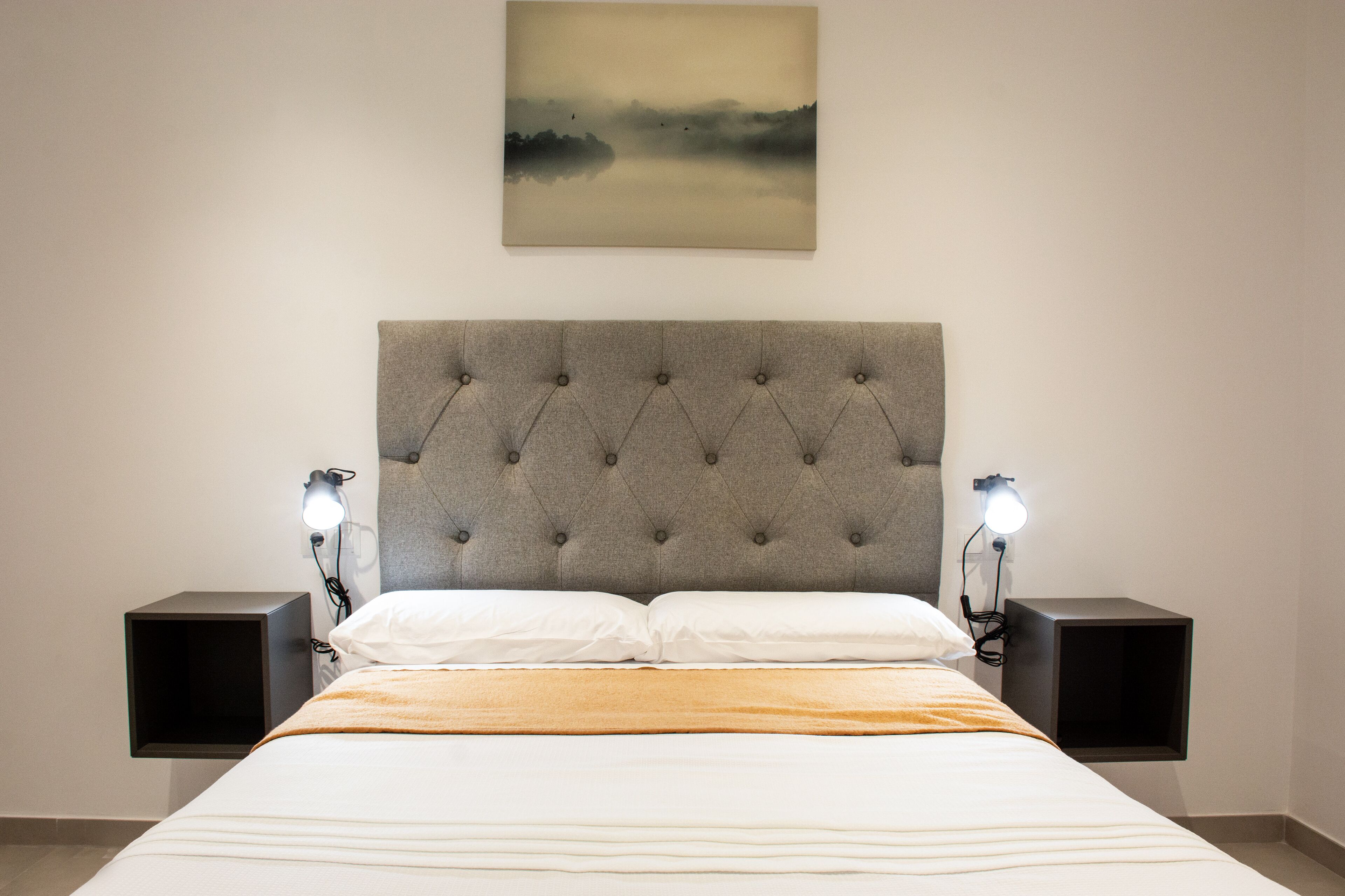 Ein ordentlich bezogenes Bett mit grauem Polsterkopfteil, zwei Nachttische mit Lampen und ein Bild an der Wand.