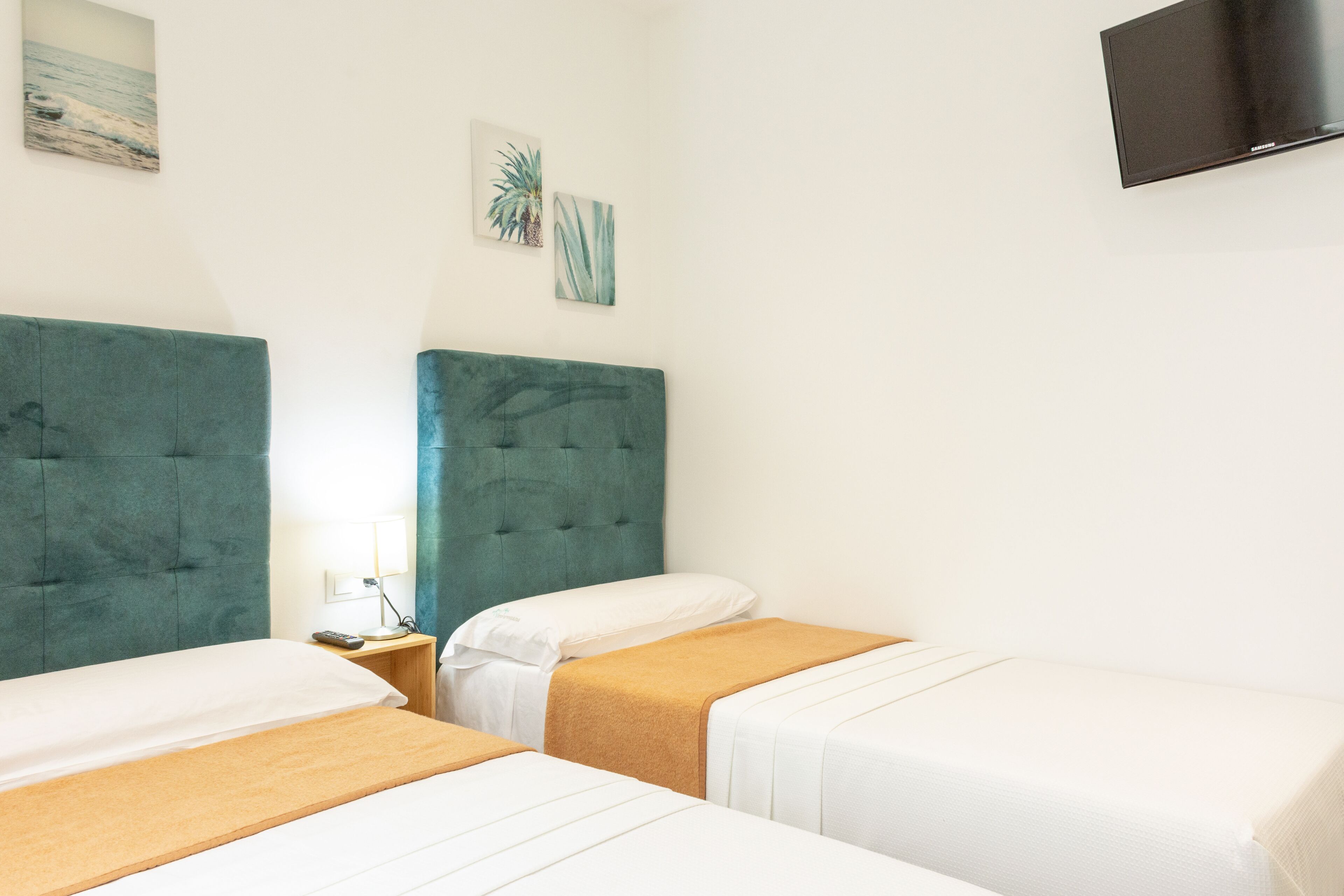 Habitación de hotel con dos camas, cada una con cabecero color turquesa y ropa de cama blanca. Hay un televisor de pantalla plana montado en la pared y una mesita de noche con una lámpara.