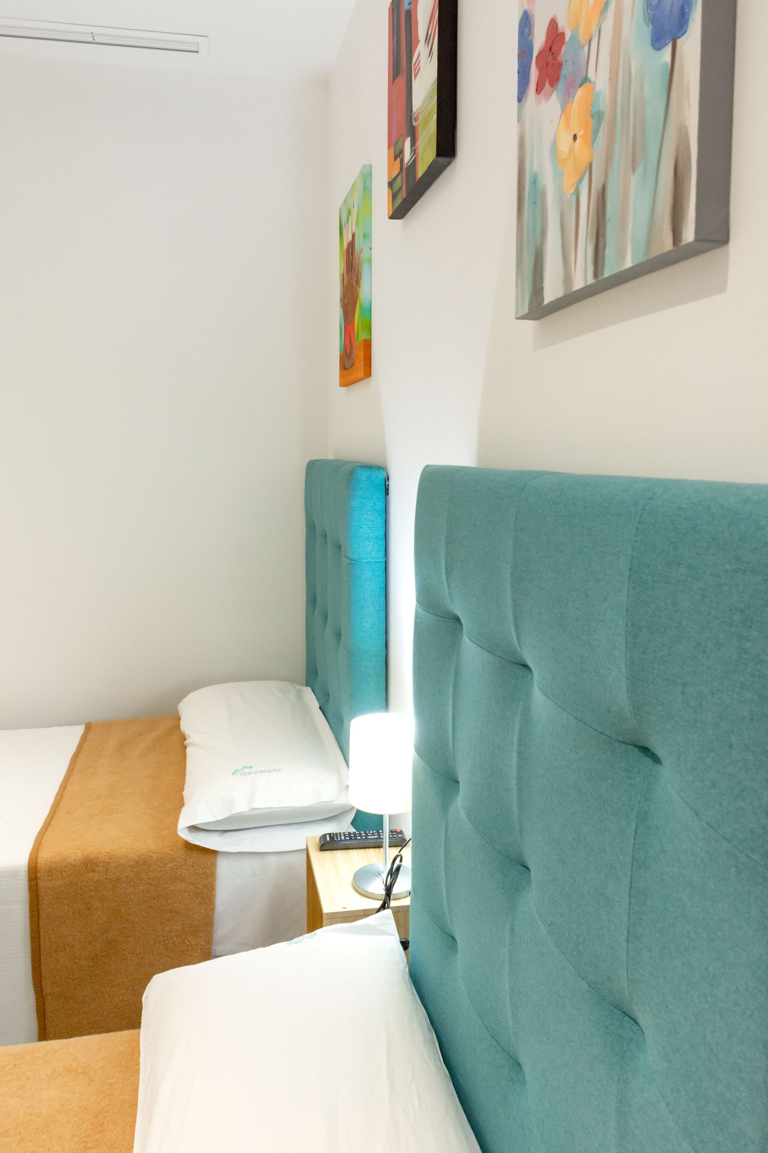 Ein Hotelzimmer mit zwei Betten, einem blauen Kopfteil, einem Nachttisch mit Lampe und einem Kunstwerk an der Wand.