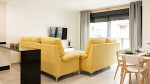 A modern living room with a yellow sofa, a dining table with chairs, and a TV on the wall. - Apartamentos Kouohana (Fuengirola)
