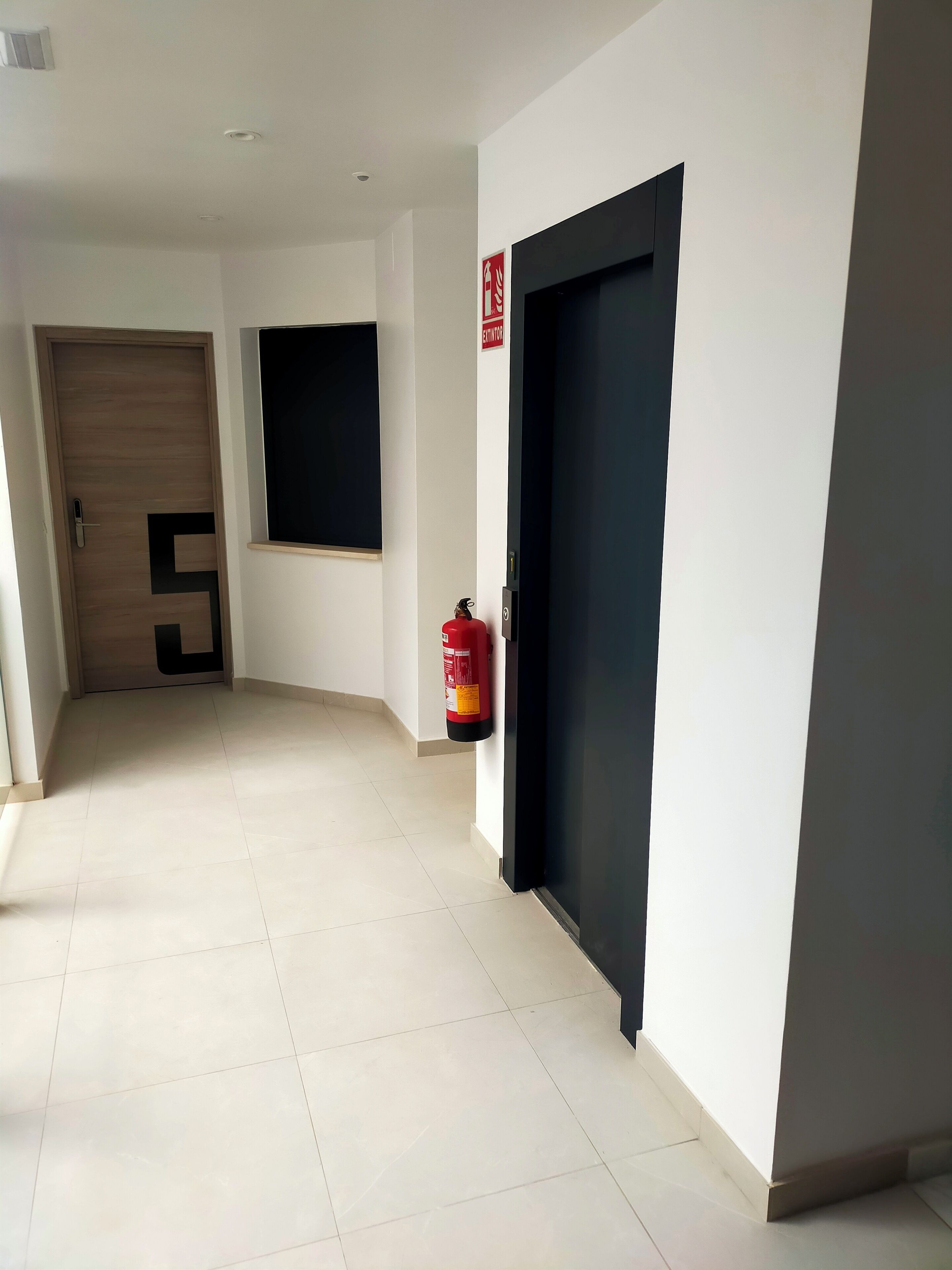 A hallway with a wooden door, a black door, a fire extinguisher, and a window.