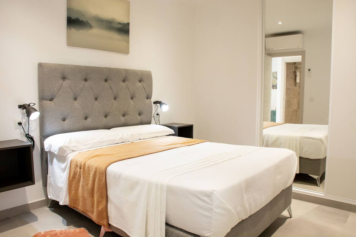 A bedroom with a large bed, two bedside tables, a wall-mounted lamp, and a painting above the bed.