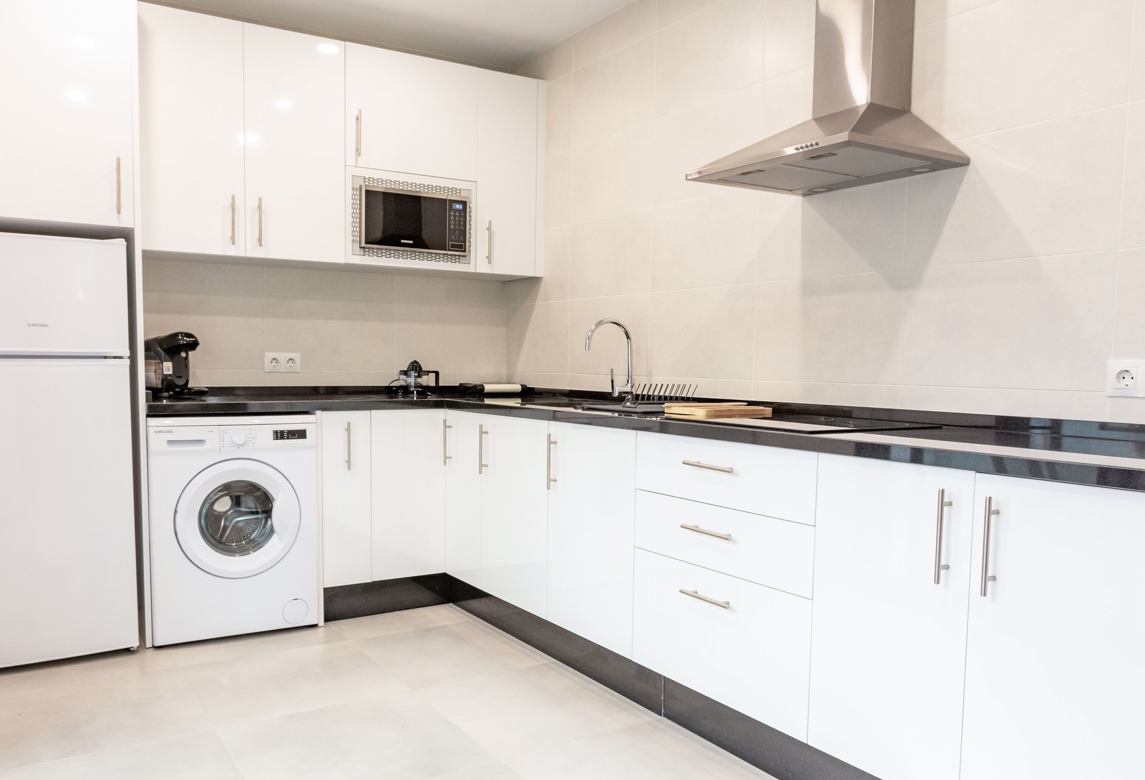 A modern kitchen with white cabinets, a built-in microwave, a stainless steel range hood, and a washing machine.