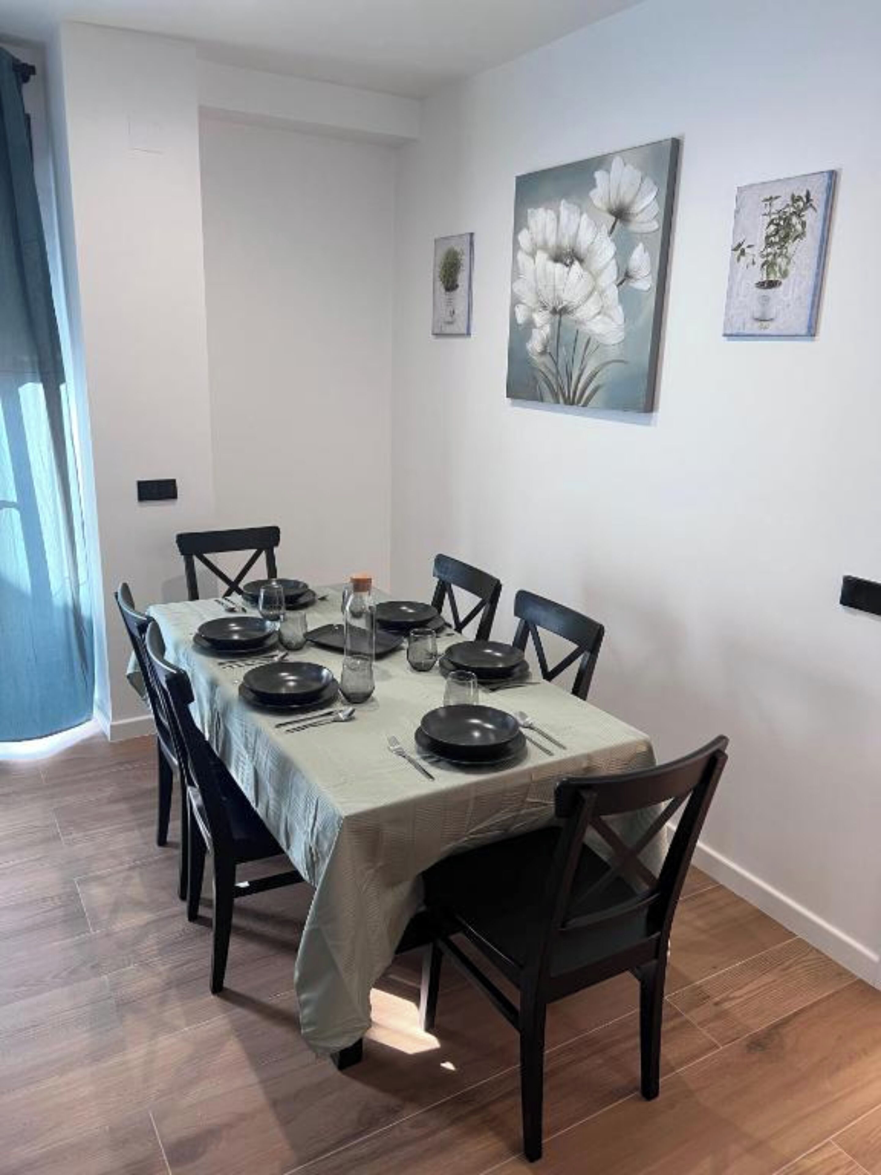 A dining area with a table set for six, surrounded by chairs, with artwork on the walls and a window with curtains.