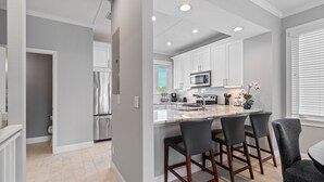 A modern kitchen with white cabinets, a stainless steel refrigerator, a microwave, and a large island with bar stools. - Pet-Friendly Cedars East Unit 847 by RVA (Longboat Key)