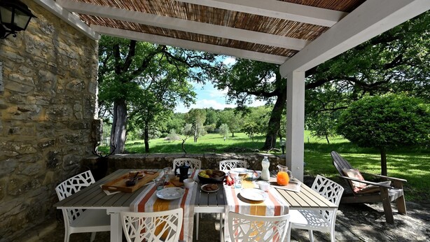 An outdoor patio with a stone wall, white lattice chairs, a wooden table set with food and drinks, and a view of a green landscape. - Antica Posta, Castellina in Chianti, Siena and Chianti (Castellina in Chianti)