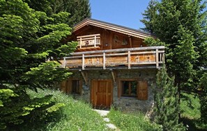 A wooden chalet with a balcony, stone foundation, and surrounded by lush greenery.