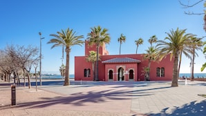 A red building with arched doorways and a tiled roof, surrounded by palm trees and a clear blue sky. - Cozy townhouse in Benalmadena 3min from the beach (Benalmádena)