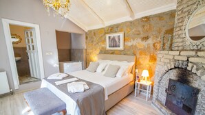 Un dormitorio con pared de piedra, chimenea, cama con almohadas, banco y una mesita de noche con lámpara.