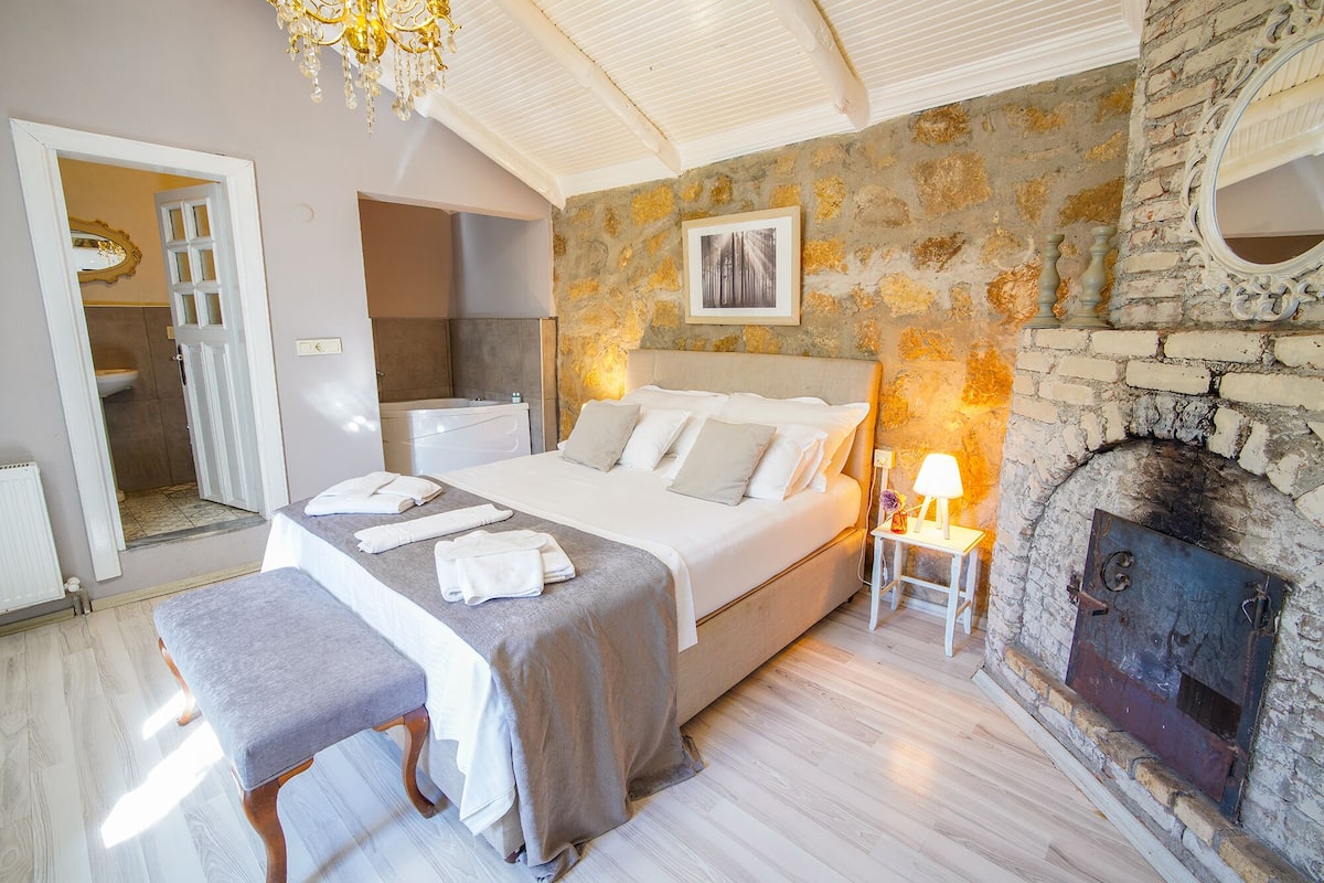 Un dormitorio con pared de piedra, chimenea, cama con almohadas, banco y una mesita de noche con lámpara.