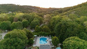 Un área de piscina con una pileta rectangular, rodeada de sillones de descanso y sombrillas, ubicada en un paisaje exuberante y boscoso con colinas al fondo.