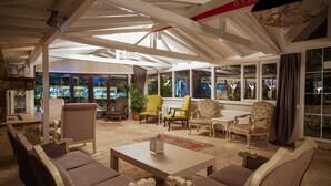 Un salón amplio con varias zonas de estar, incluyendo sofás y sillones, dispuestos alrededor de una mesa de centro. Amplias ventanas ofrecen vistas al exterior.
