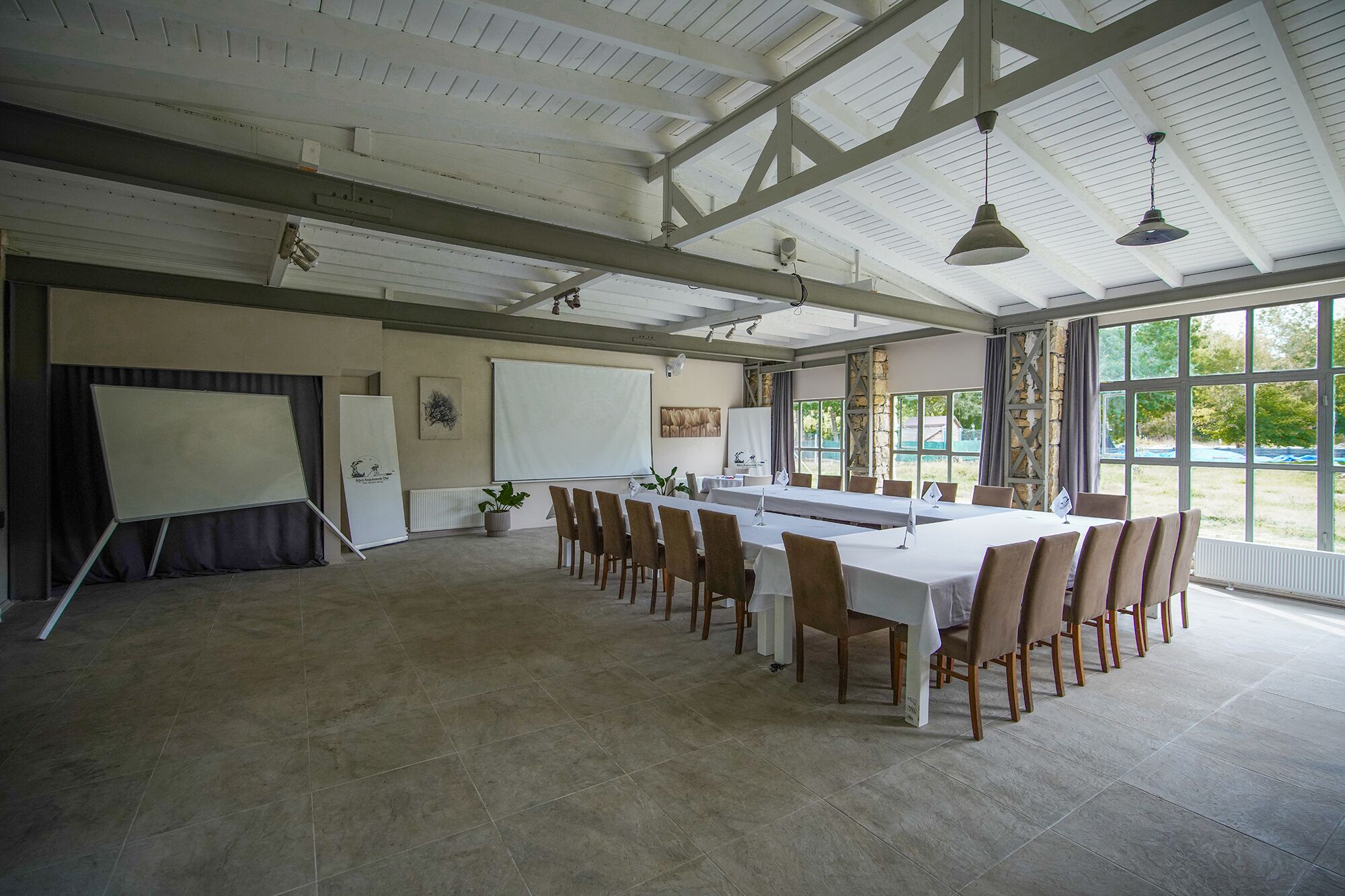 Uma sala de conferências ampla com uma mesa comprida, cadeiras, uma tela de projetor e uma grande janela com vista para vegetação.