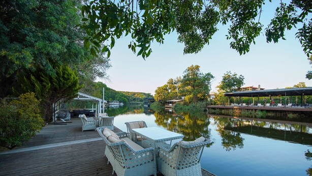 Una terraza de madera con mobiliario de mimbre y una mesa, con vista a un lago tranquilo y edificios al fondo.