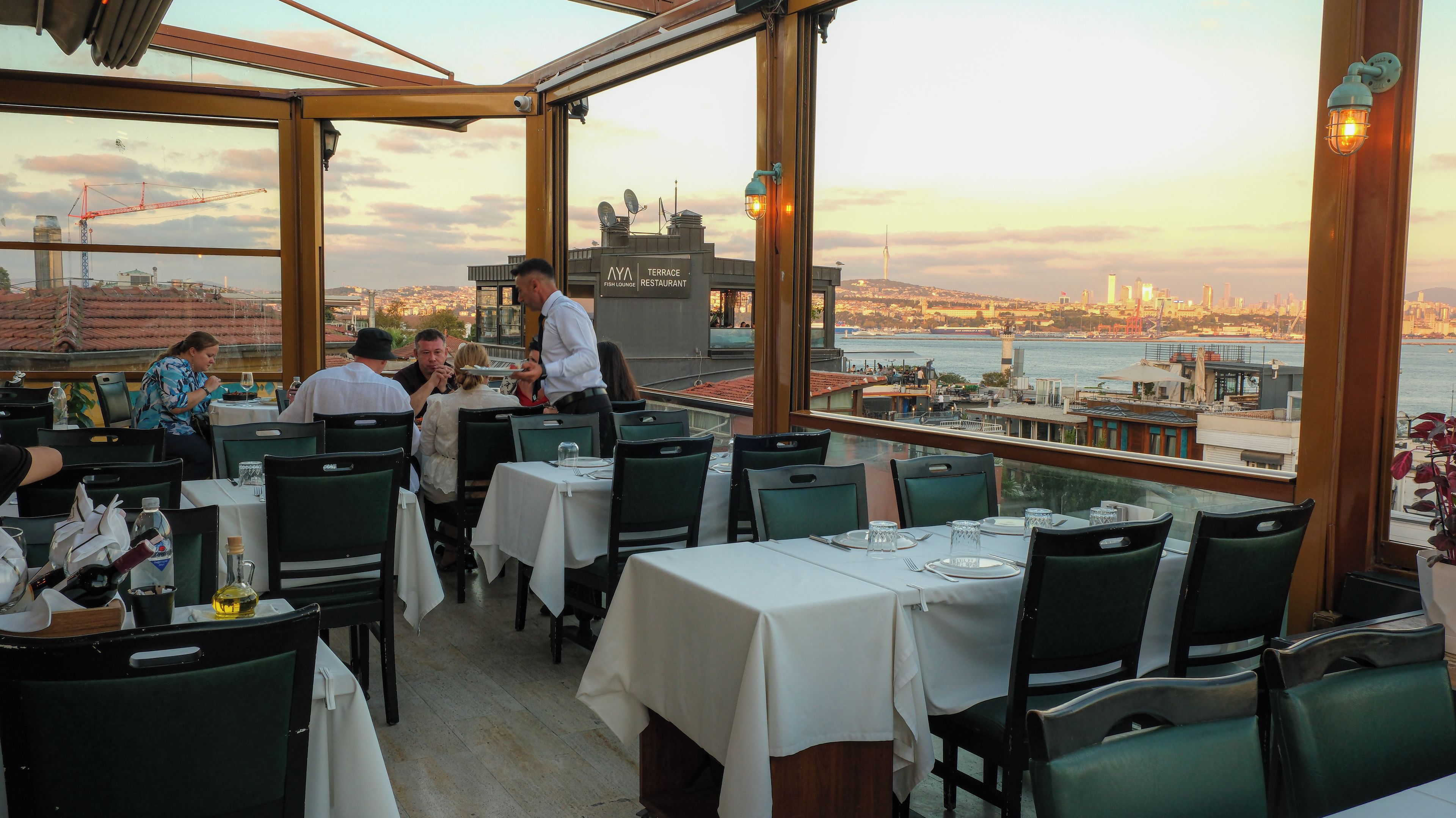 A restaurant with tables set for dining, a view of the cityscape, and a construction crane in the background.