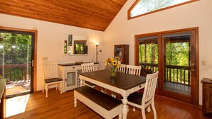 Un espace repas avec une table en bois, des chaises blanches et un banc. Le plafond est en bois, il y a une fenêtre et une porte qui donne sur une terrasse extérieure.