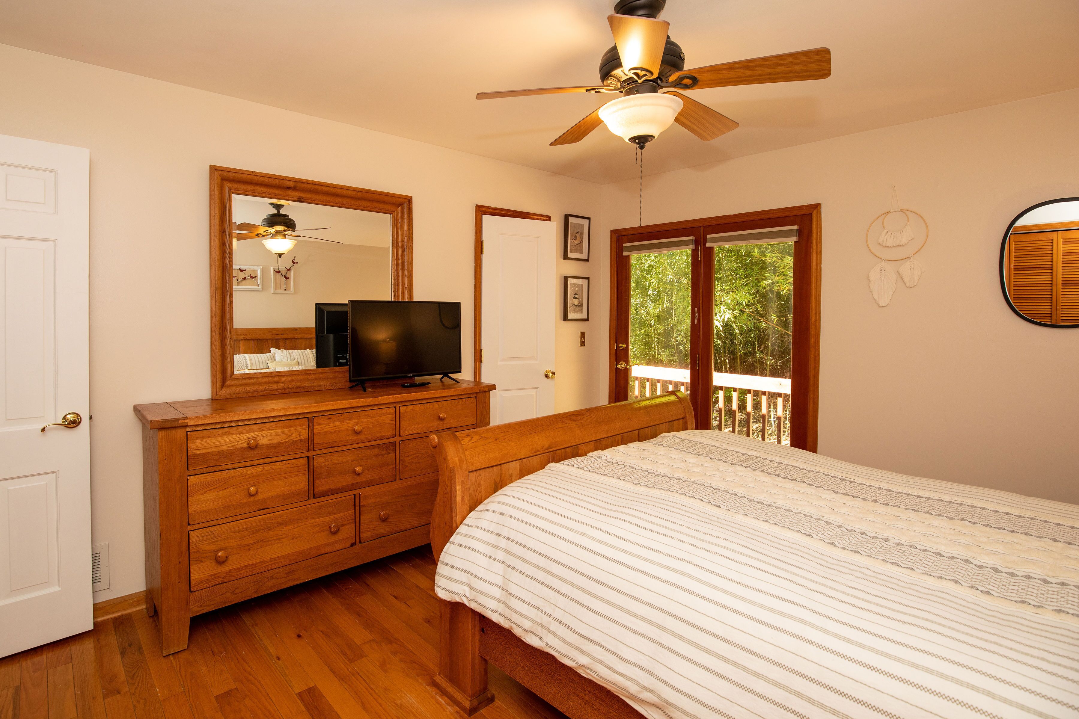 A bedroom with a wooden dresser, a television, a ceiling fan, and a large window with a view of greenery.