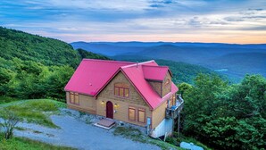 A wooden cabin with a red roof, surrounded by lush greenery and mountains in the background. - Gorgeous Chalet - Incredible Views - Hot Tub - Fire Pit (Snowshoe)