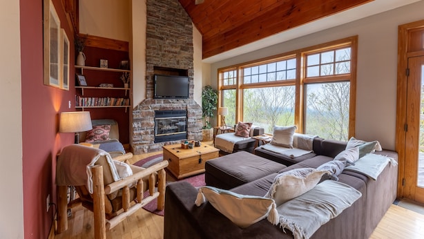 A cozy living room with a stone fireplace, wooden ceiling, and large windows offering a view of the outdoors. - Gorgeous Chalet - Incredible Views - Hot Tub - Fire Pit (Snowshoe)