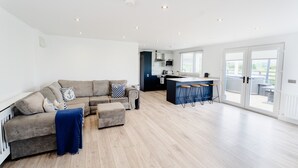 A modern living room with a sofa, ottoman, and a blue throw blanket. A kitchen with a bar area is visible in the background. - Castle View Chalet by Clonlum Cottages (Newry)