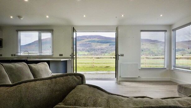 A modern living room with a sofa, a kitchen area, and large windows offering a view of the countryside. - Castle View Chalet by Clonlum Cottages (Newry)