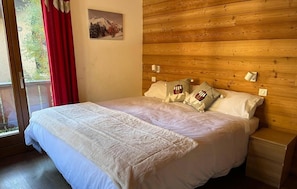 A bedroom with a wooden headboard, a bed with white bedding, two pillows with a mountain design, and a window with red curtains.